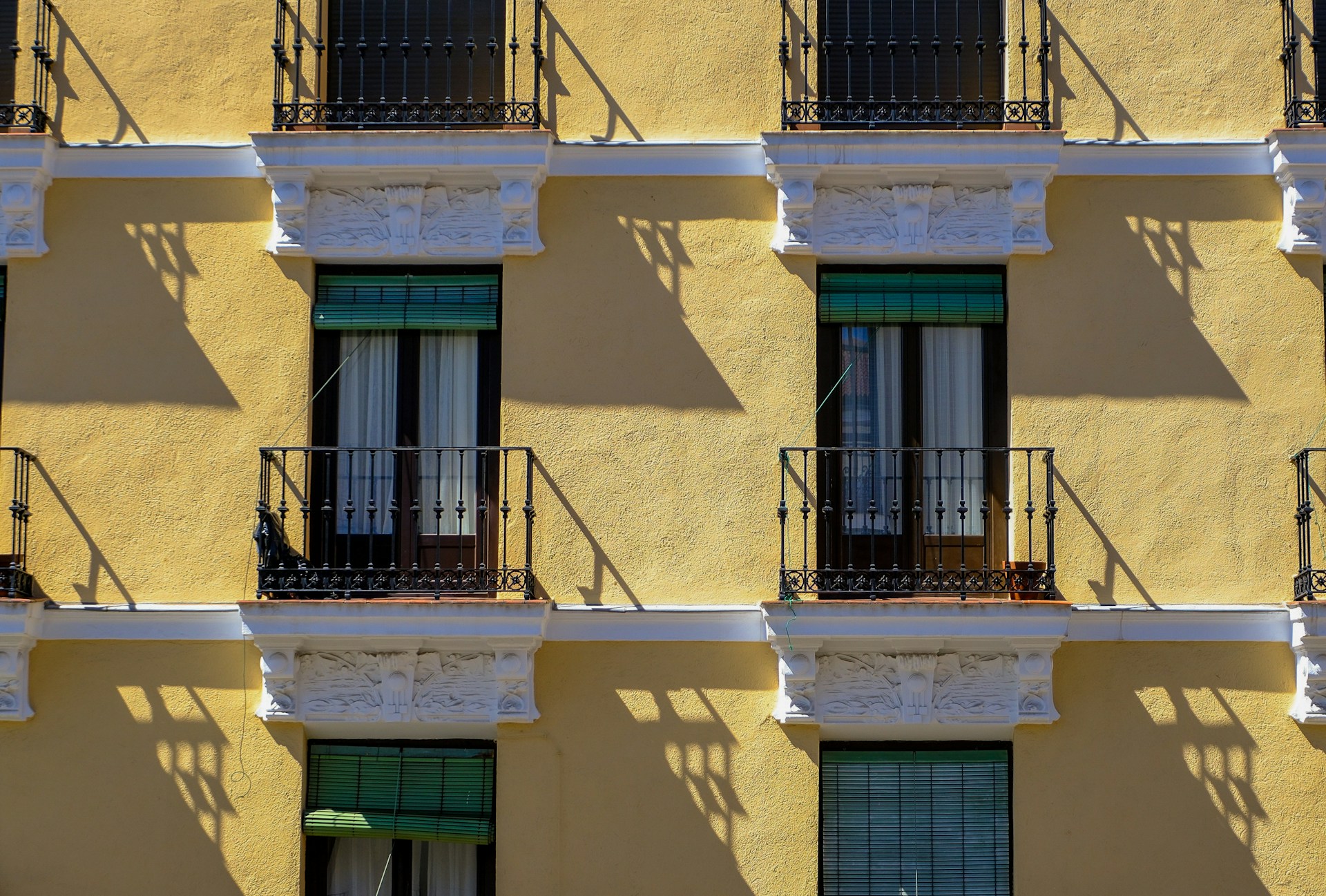 Fachada amarilla con balcones de hierro, macetas de barro y persianas verdes, bajo un sol duro que proyecta sombras nítidas.
