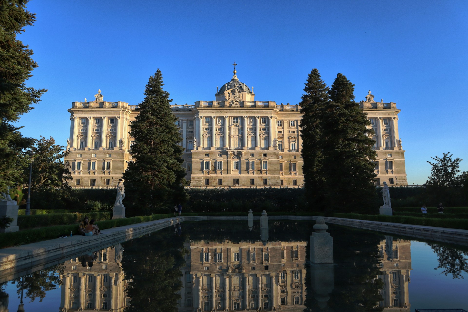 Luz cálida de tarde en los Jardines de Sabatini de Madrid: el Palacio Real, el estanque en calma y dos personas sentadas al borde.