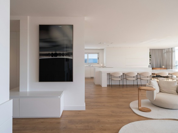 Interior diáfano de apartamento con isla blanca, taburetes, sofá claro y suelo de madera, con luz natural desde grandes ventanales