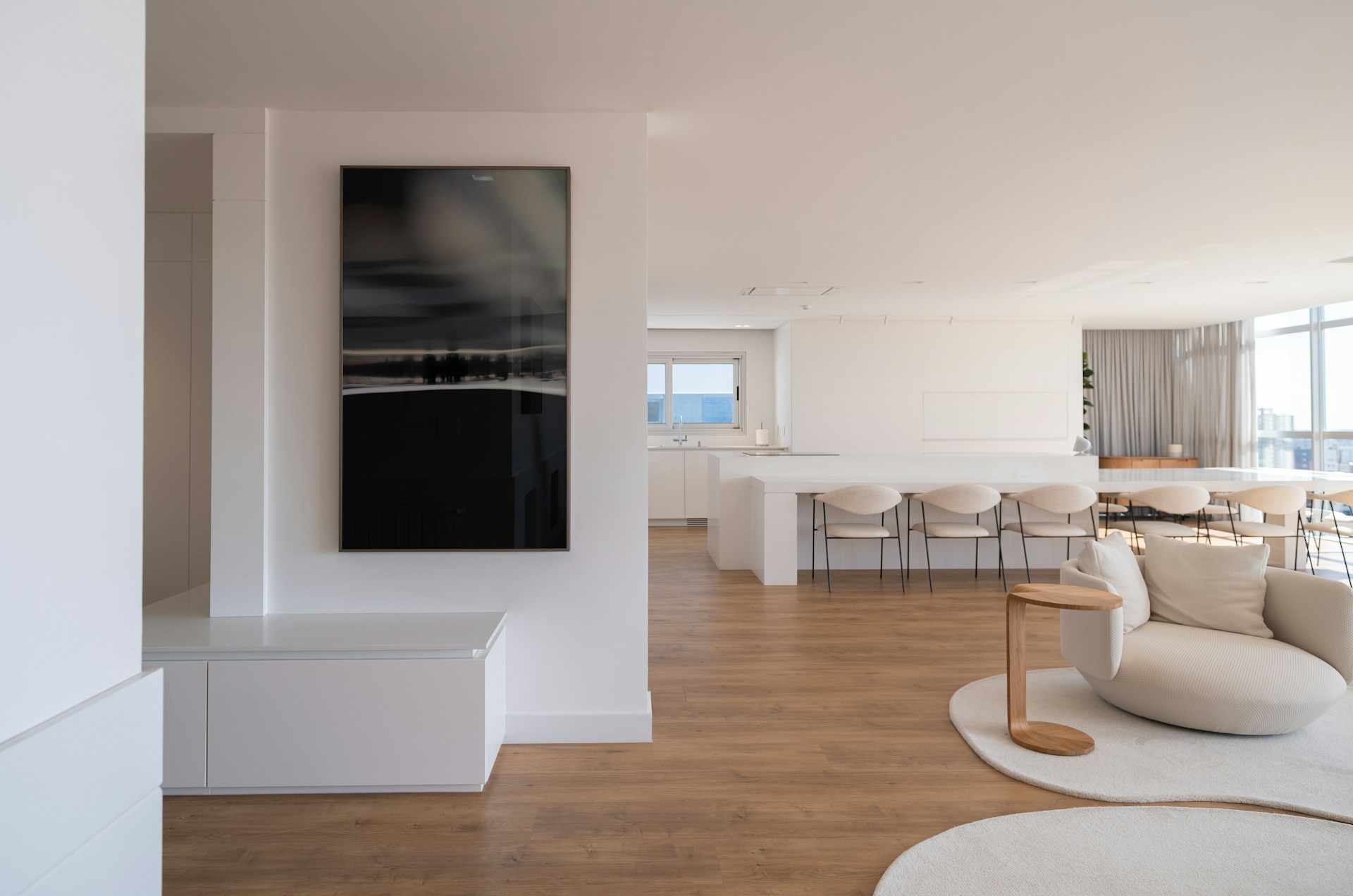 Interior diáfano de apartamento con isla blanca, taburetes, sofá claro y suelo de madera, con luz natural desde grandes ventanales