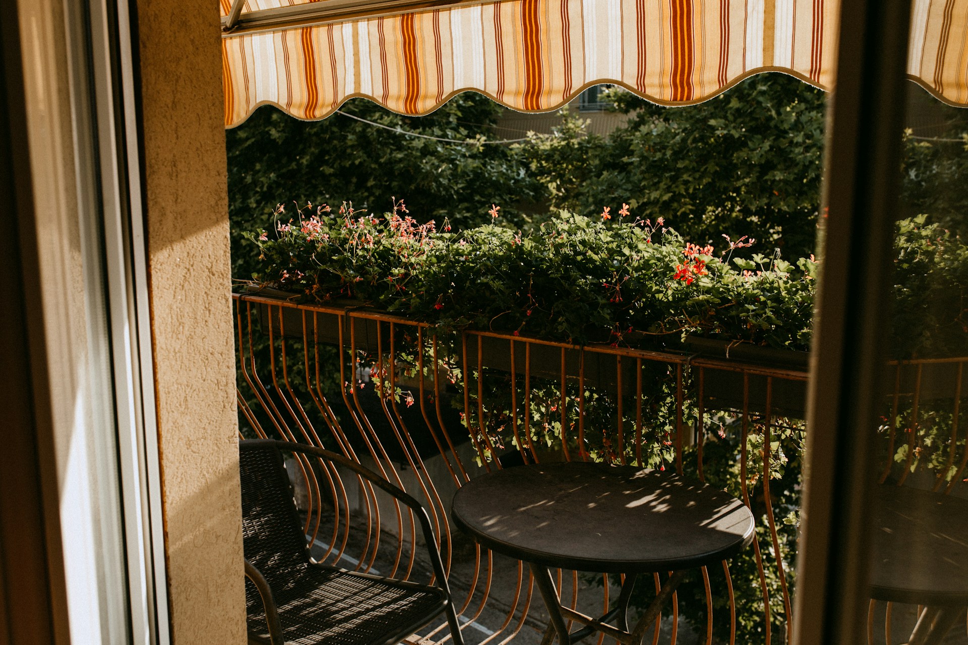 Balcón con toldo de rayas, barandilla metálica y jardineras de geranios en flor, con una mesa redonda y una silla bajo la sombra moteada de los árboles.