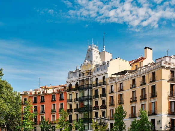 Fachadas residenciales del siglo XIX en el centro de Madrid, con balcones de hierro, un mirador acristalado y copas de árboles verdes en primer plano.