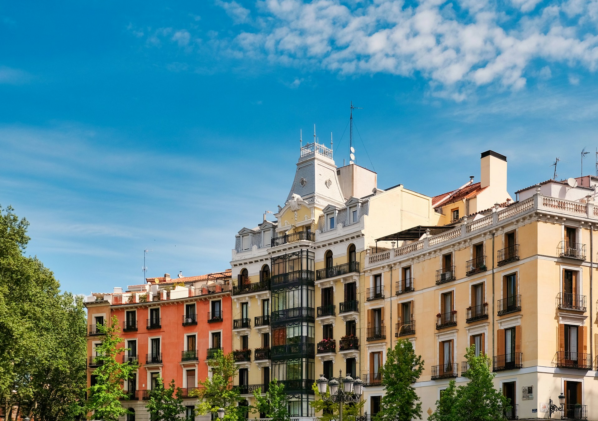 Fachadas residenciales del siglo XIX en el centro de Madrid, con balcones de hierro, un mirador acristalado y copas de árboles verdes en primer plano.