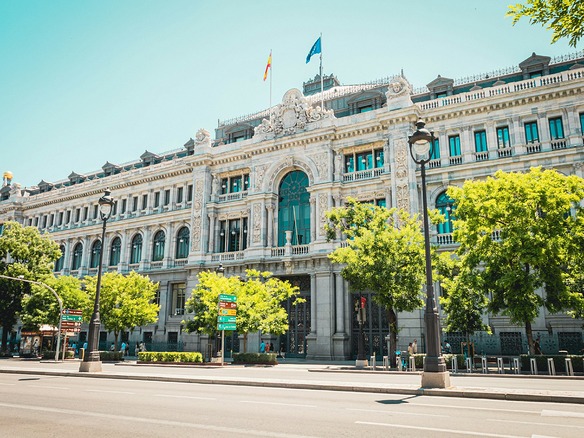 Calzada vacía de la Calle de Alcalá en Madrid, con la fachada del Banco de España, arbolado verde y algunos peatones en la acera bajo el sol de verano.