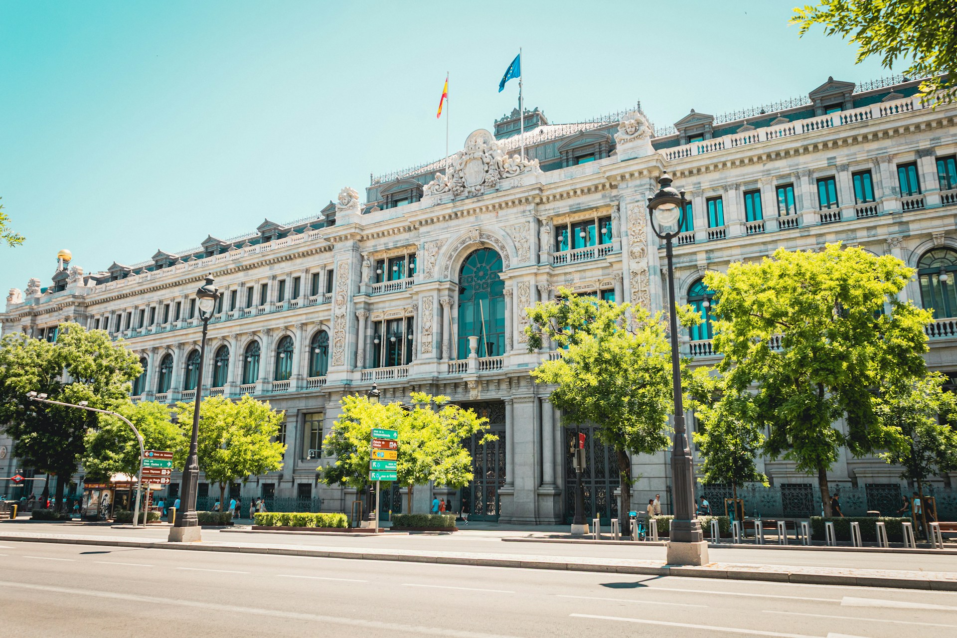 Calzada vacía de la Calle de Alcalá en Madrid, con la fachada del Banco de España, arbolado verde y algunos peatones en la acera bajo el sol de verano.