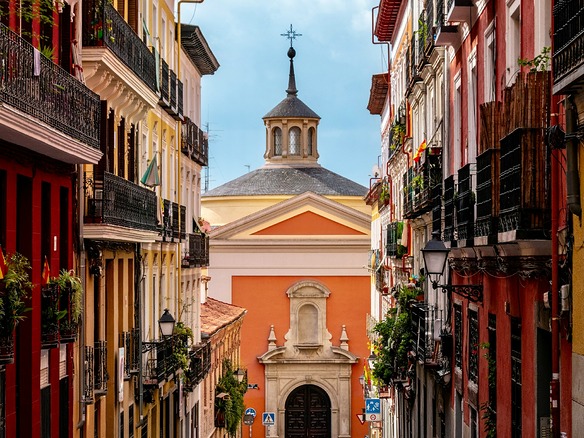 Calle estrecha y vacía entre fachadas rojas y amarillas con balcones de forja, cerrada al fondo por una iglesia de muros anaranjados y cúpula gris.