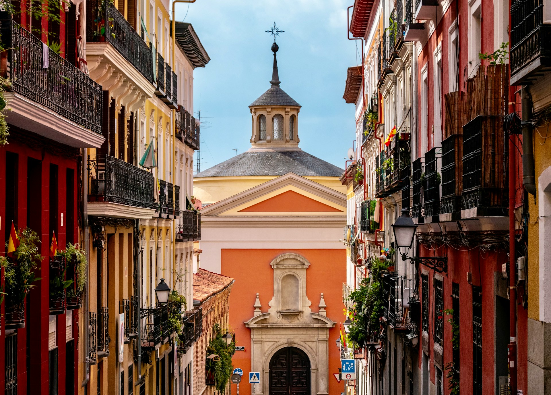 Calle estrecha y vacía entre fachadas rojas y amarillas con balcones de forja, cerrada al fondo por una iglesia de muros anaranjados y cúpula gris.