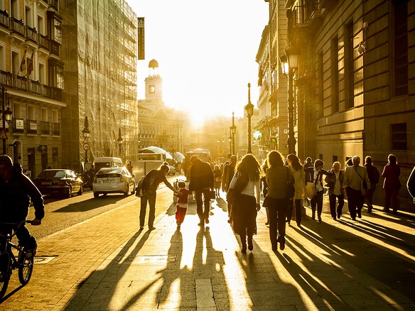 Peatones caminando a contraluz por una calle del centro de Madrid al atardecer, con la torre del reloj de la Puerta del Sol al fondo