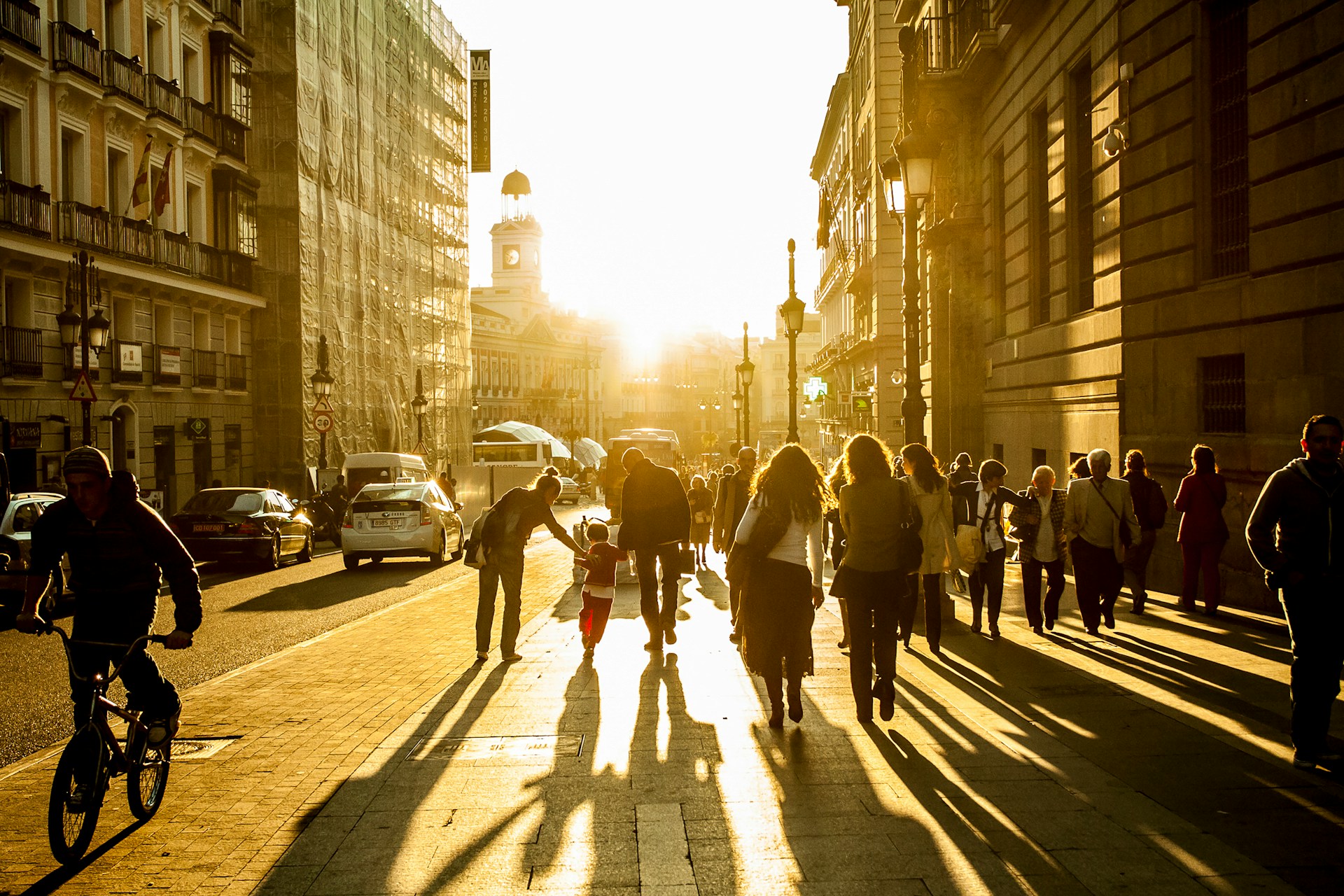 Peatones caminando a contraluz por una calle del centro de Madrid al atardecer, con la torre del reloj de la Puerta del Sol al fondo