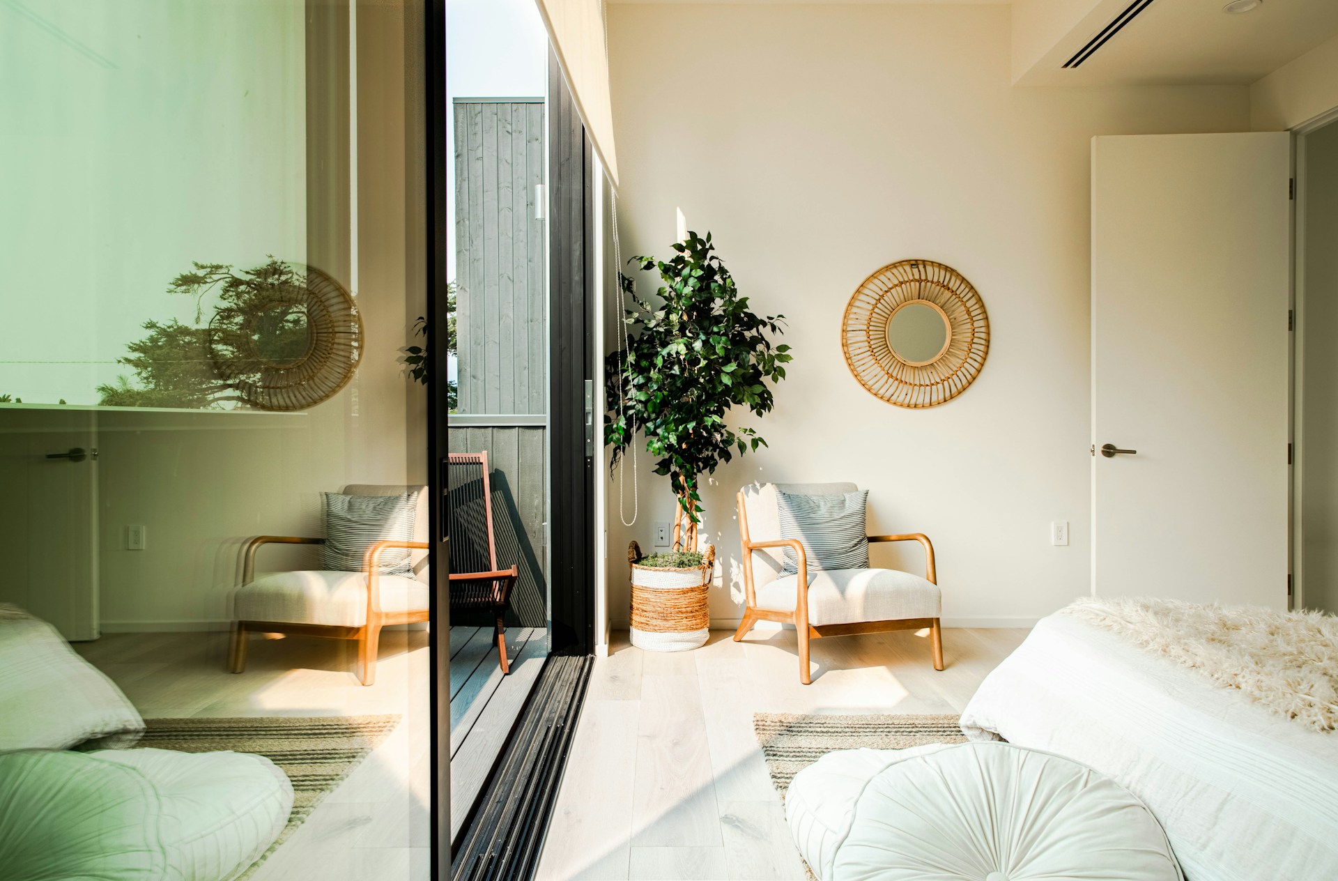 Estancia luminosa con butaca de madera y tela, espejo redondo de ratán, planta de interior y puerta corredera abierta a una terraza de madera con una silla.
