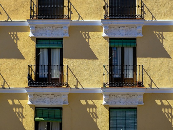 Fachada amarilla con balcones de hierro, macetas de barro y persianas verdes, bajo un sol duro que proyecta sombras nítidas.