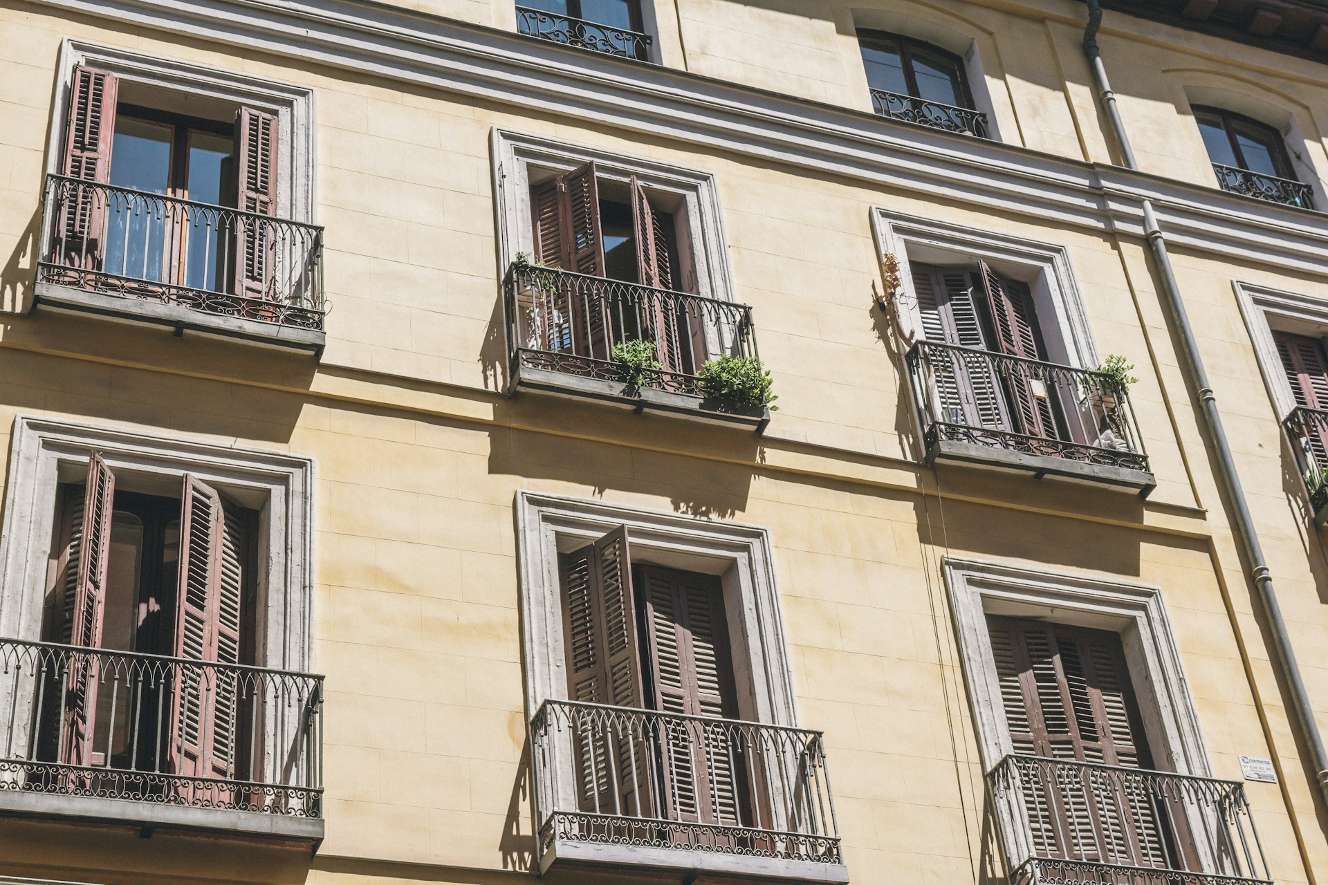 Fachada color crema de un edificio de viviendas de Madrid con balcones de forja, contraventanas de madera y macetas, bajo el sol duro del verano.