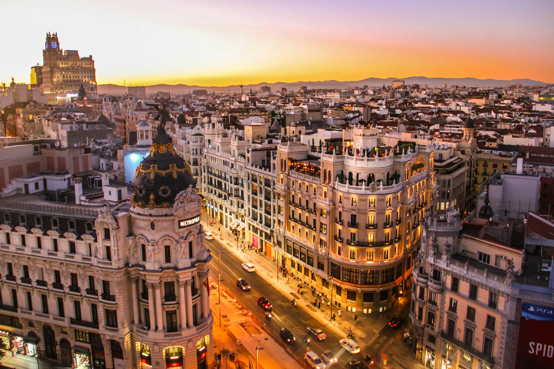 Vista elevada de la Gran Vía de Madrid al atardecer, con el edificio Metrópolis, terrazas en las azoteas y fachadas iluminadas