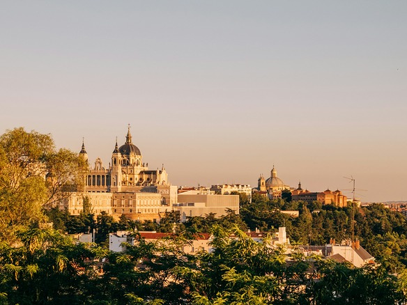 Vista de Madrid al atardecer: la cúpula de la catedral de la Almudena y el Palacio Real en piedra clara, bañados de luz dorada sobre copas de árboles verdes y tejados.