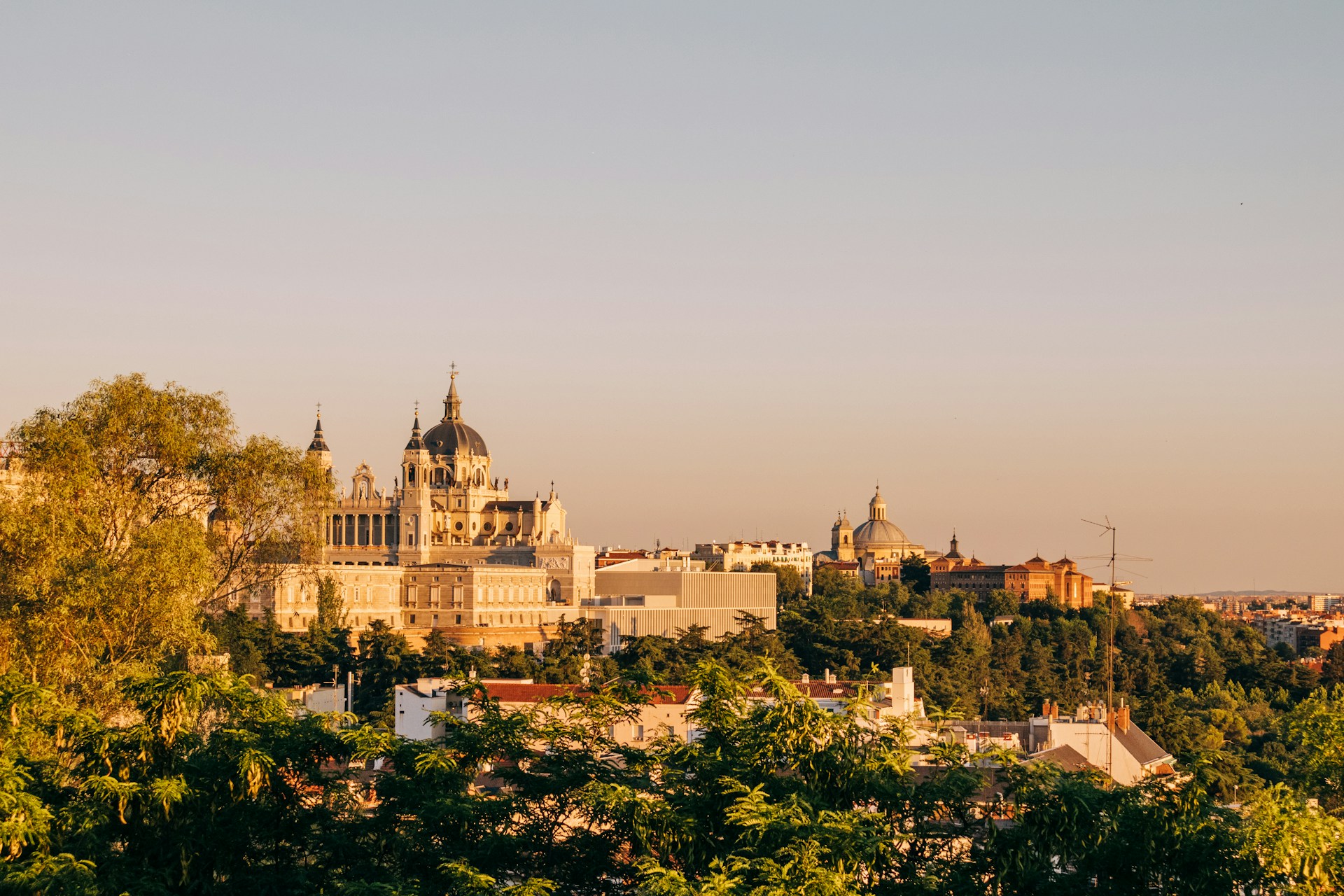 Vista de Madrid al atardecer: la cúpula de la catedral de la Almudena y el Palacio Real en piedra clara, bañados de luz dorada sobre copas de árboles verdes y tejados.