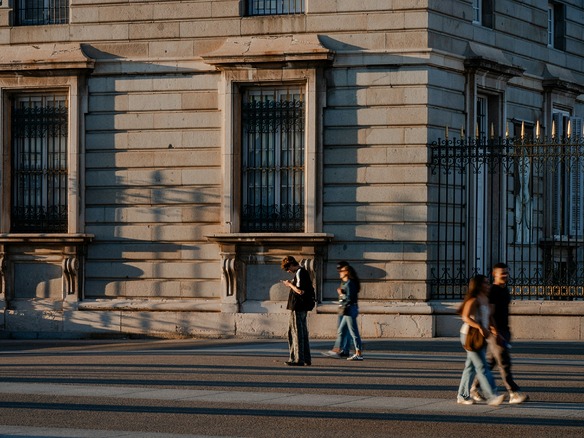 Fachada de piedra de un palacio de Madrid bajo luz cálida y rasante, con verja de hierro de remates dorados y transeúntes en la explanada.