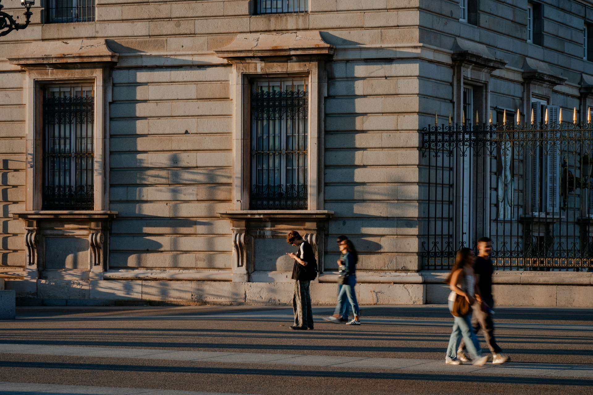 Fachada de piedra de un palacio de Madrid bajo luz cálida y rasante, con verja de hierro de remates dorados y transeúntes en la explanada.
