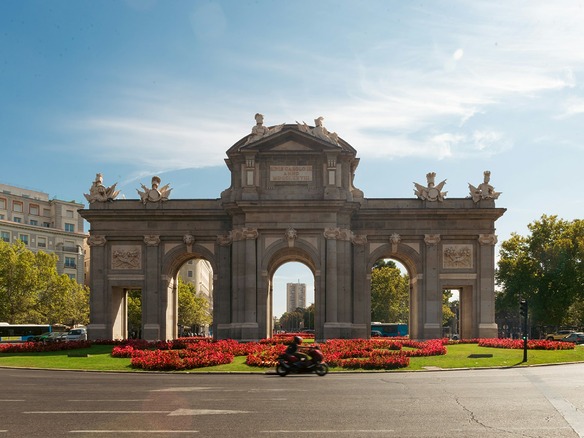 Arco de piedra de la Puerta de Alcalá en Madrid, rodeado de parterres de flores rojas y árboles verdes, con un motorista solitario cruzando la amplia calzada casi vacía.
