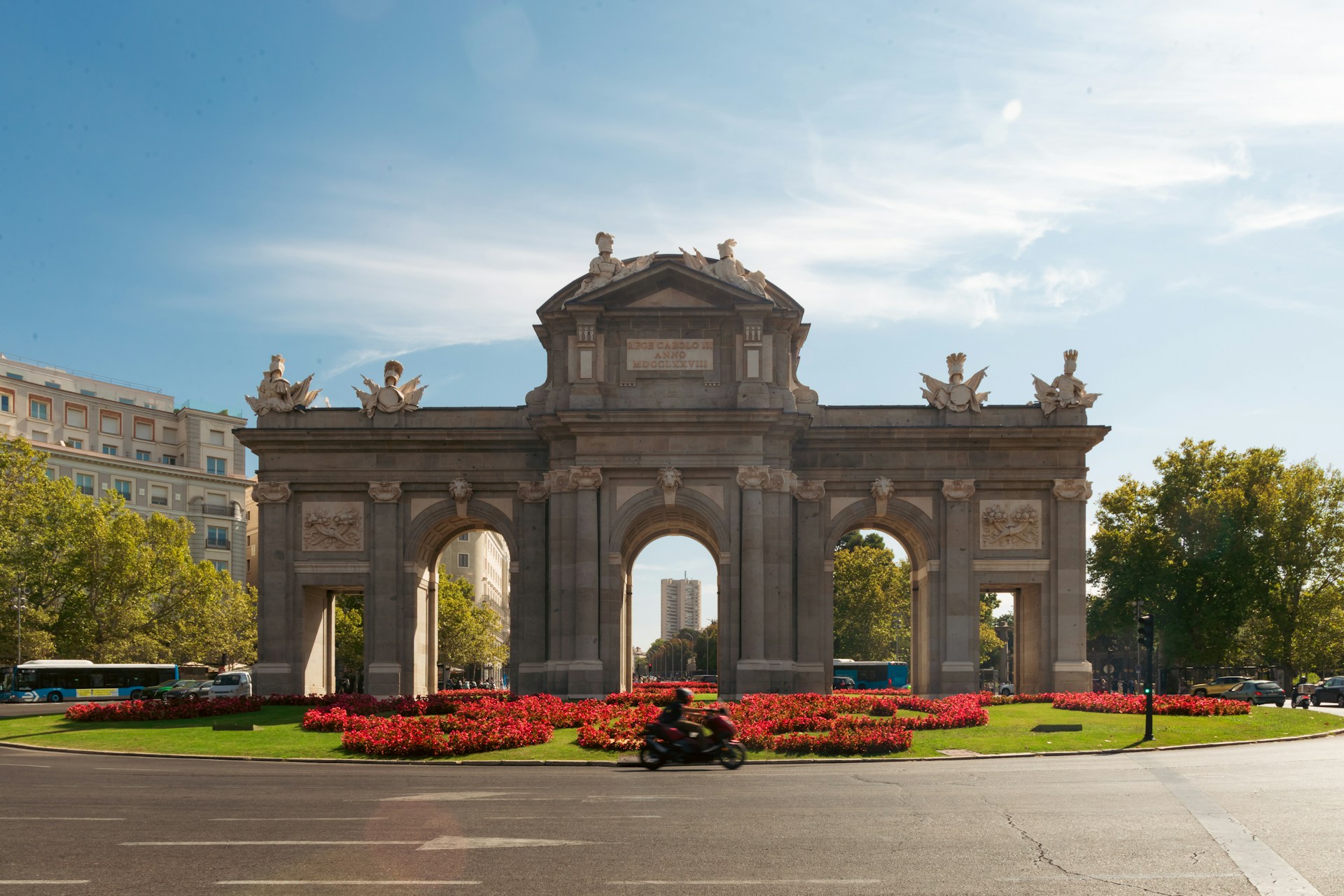 Arco de piedra de la Puerta de Alcalá en Madrid, rodeado de parterres de flores rojas y árboles verdes, con un motorista solitario cruzando la amplia calzada casi vacía.