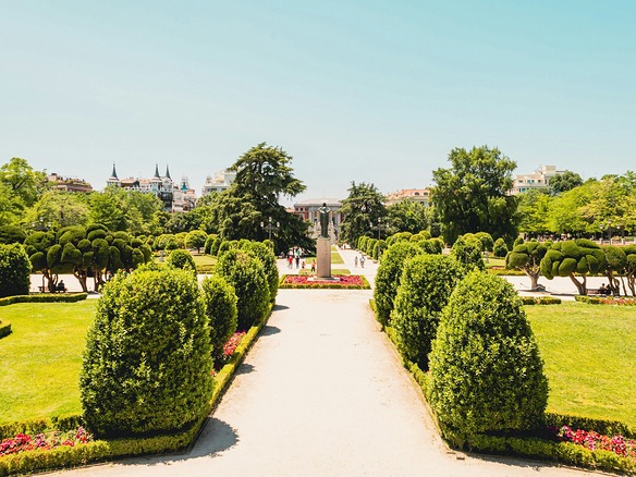 Jardín del Parque de El Retiro en Madrid: setos cónicos recortados y cipreses podados, césped verde y flores rojas junto a un ancho paseo soleado, con algunos paseantes a lo lejos.