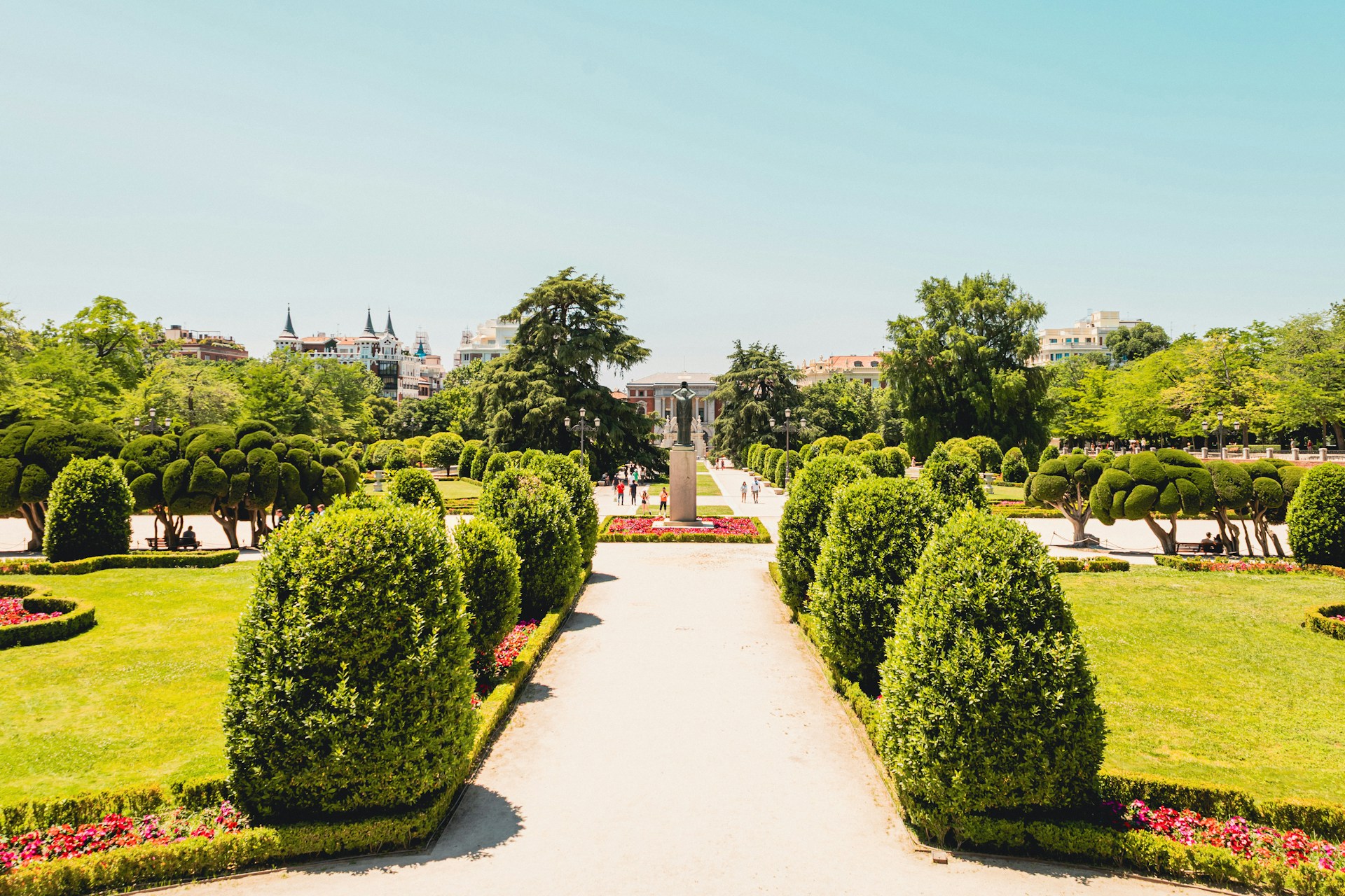 Jardín del Parque de El Retiro en Madrid: setos cónicos recortados y cipreses podados, césped verde y flores rojas junto a un ancho paseo soleado, con algunos paseantes a lo lejos.