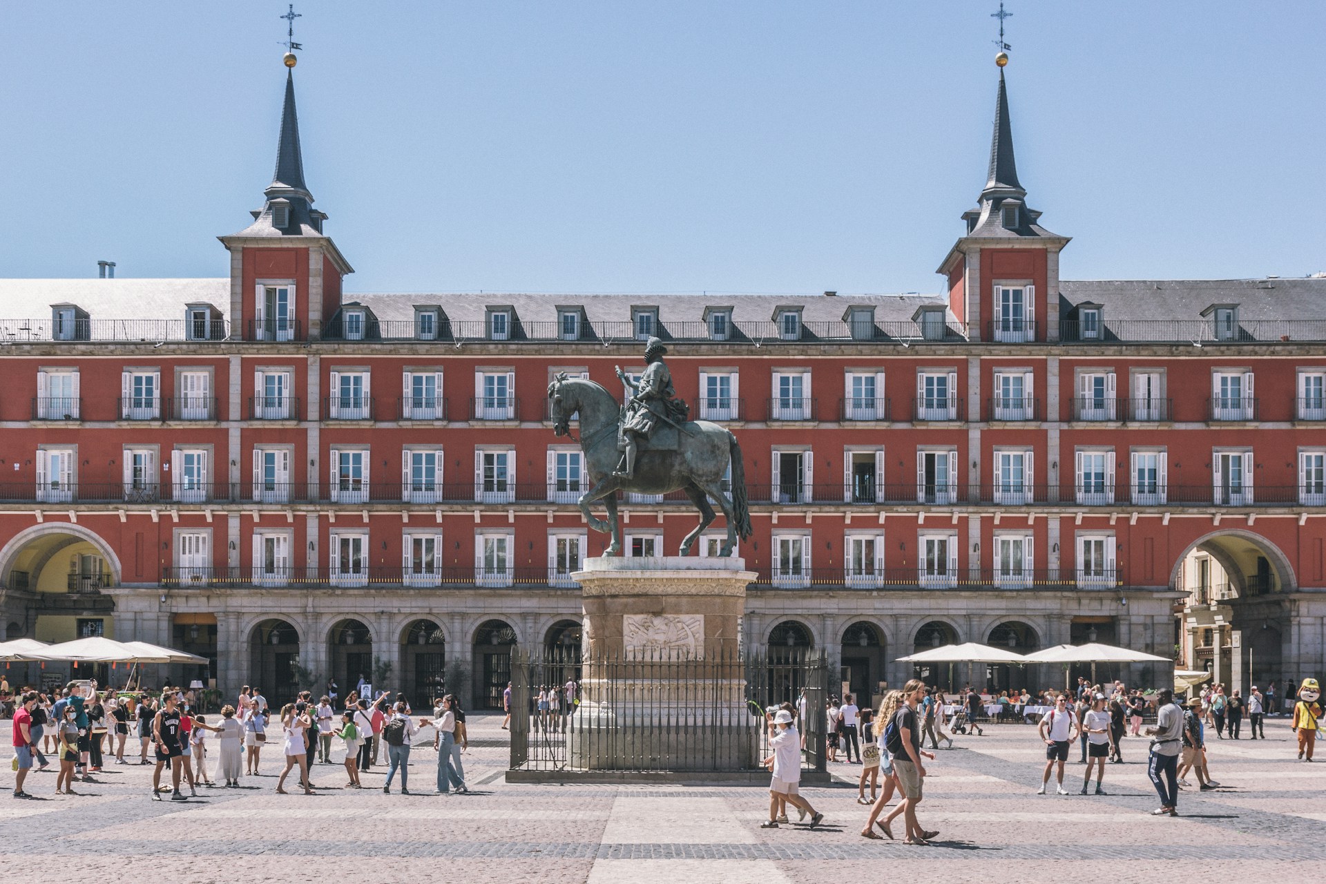 Plaza Mayor de Madrid en un día despejado, con la estatua ecuestre, los soportales y las sombrillas blancas de las terrazas llenas de gente.