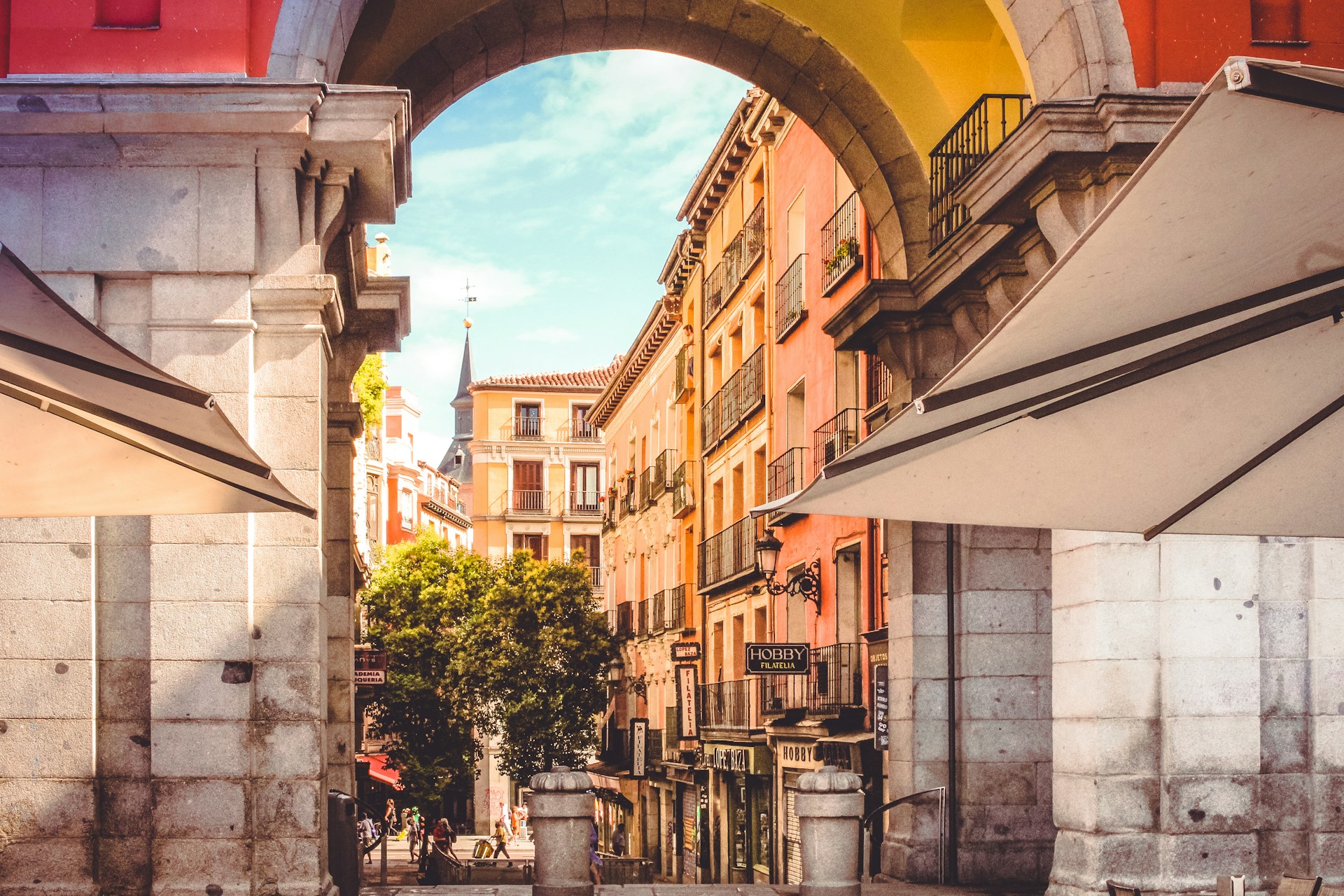 Fachadas ocres y balcones de hierro vistos desde un arco de la Plaza Mayor de Madrid, con toldos y sombrillas dando sombra.