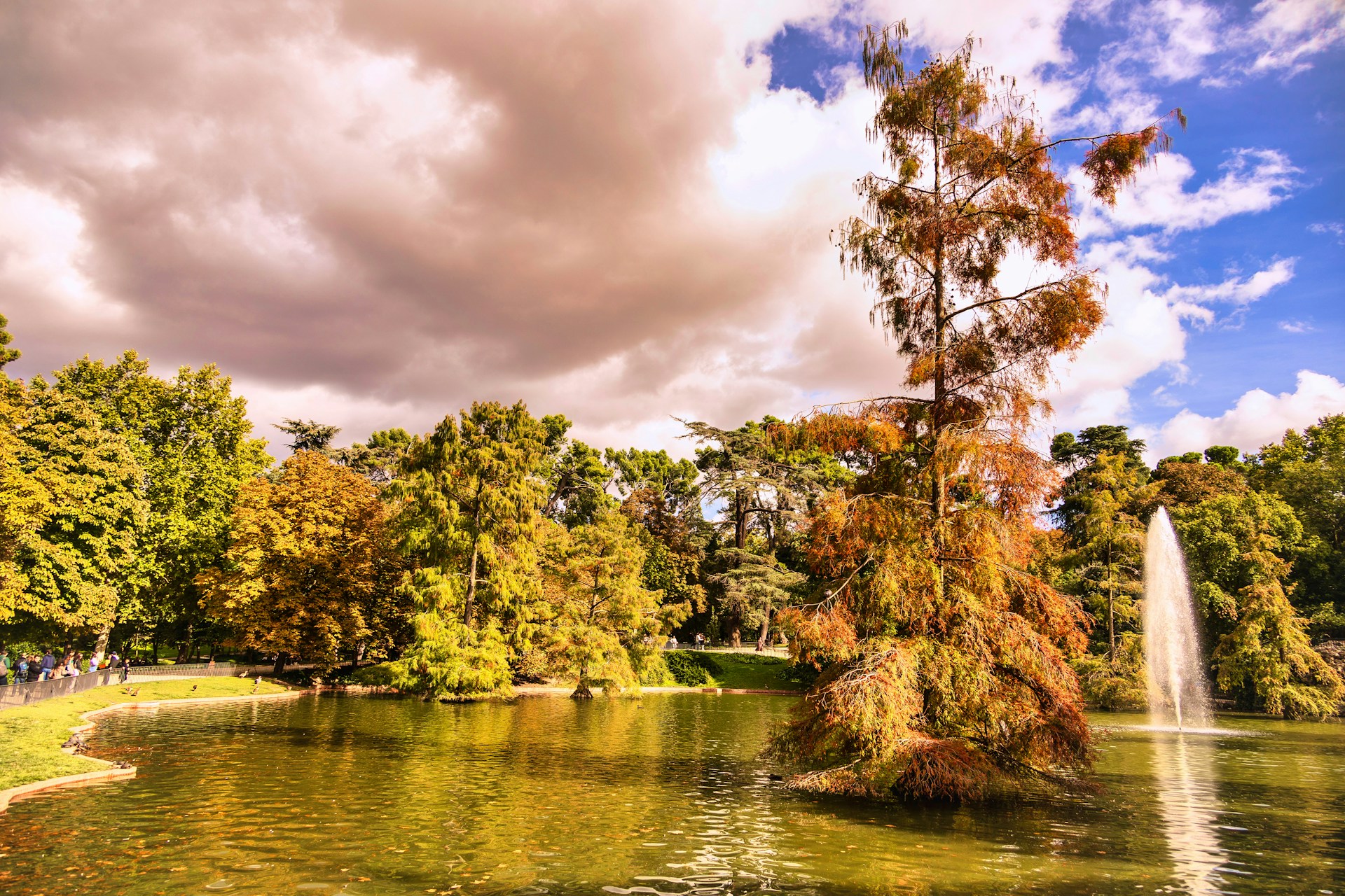 Estanque del parque del Retiro en Madrid con cipreses calvos cobrizos, un surtidor de agua y árboles dorados bajo un cielo nuboso de octubre.