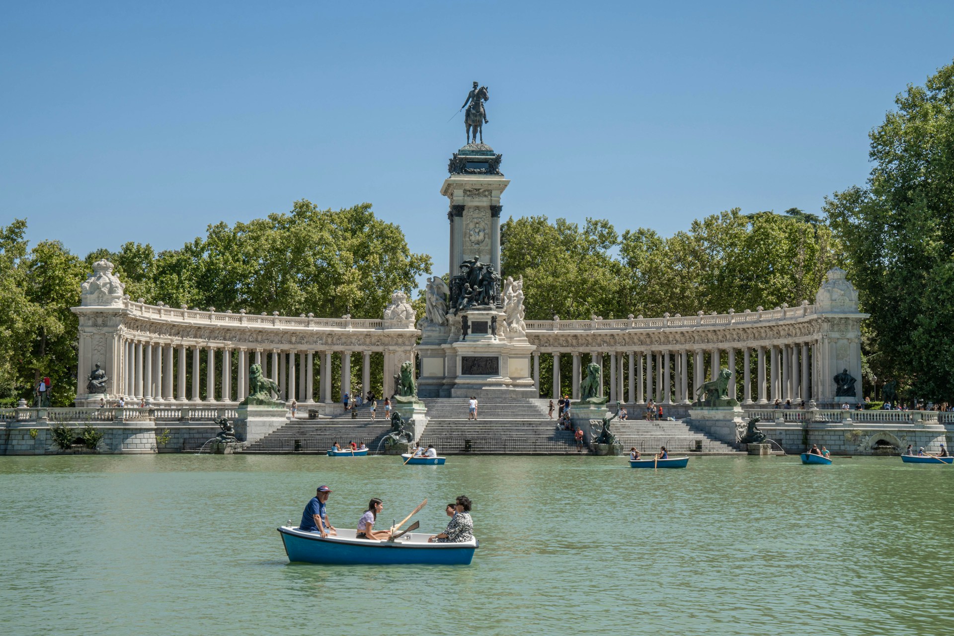Barcas de remos azules en el estanque del Retiro frente a la columnata del monumento a Alfonso XII, entre árboles verdes y cielo azul.